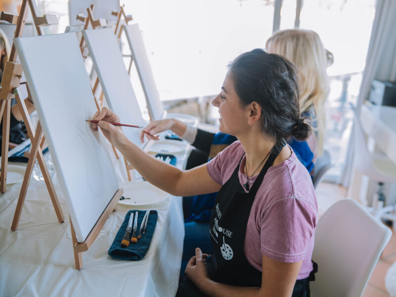 A smiling woman drawing an outline on a paint canvas.